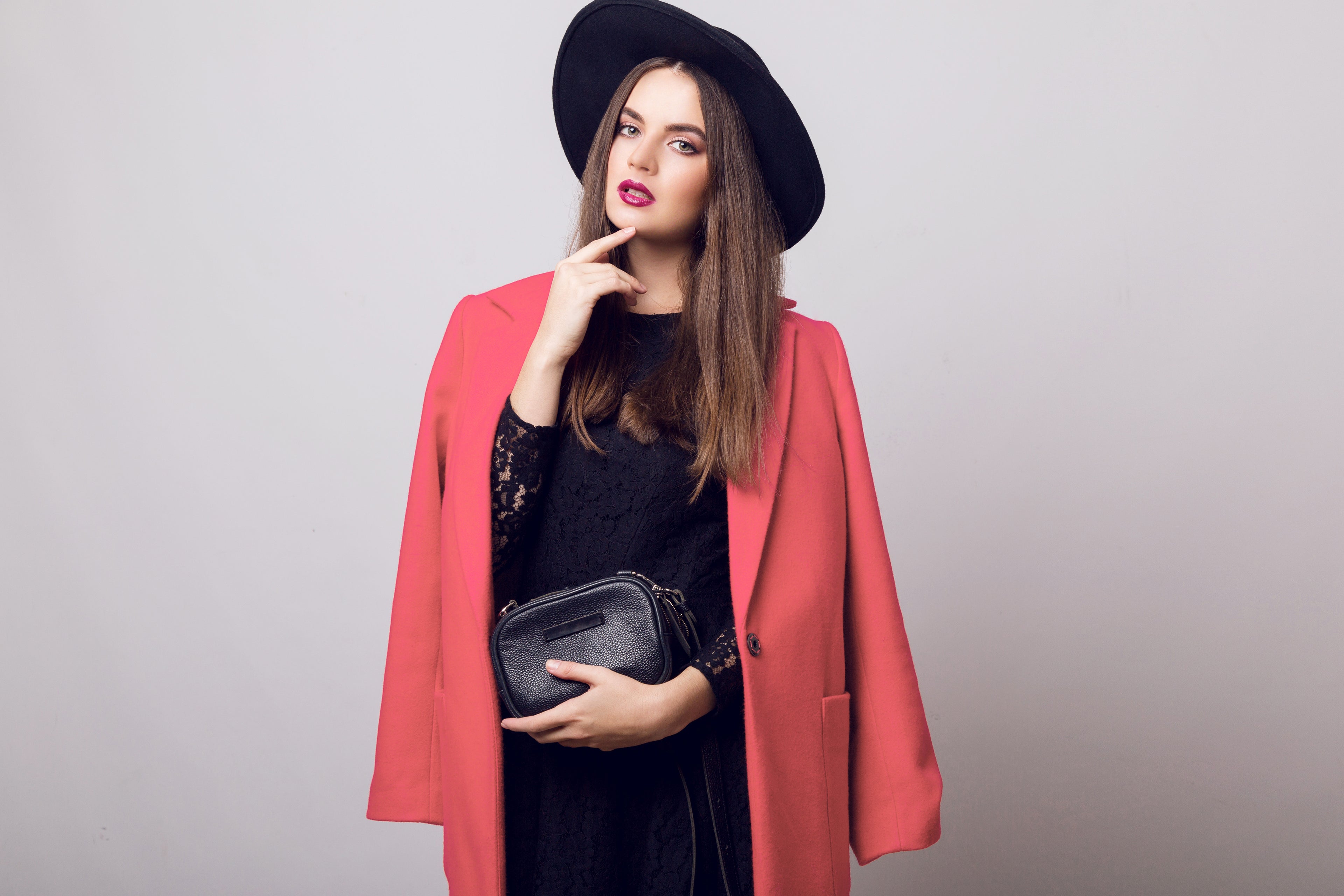 Fashionable woman in a pink cape coat and black hat, holding a handbag, posing elegantly.